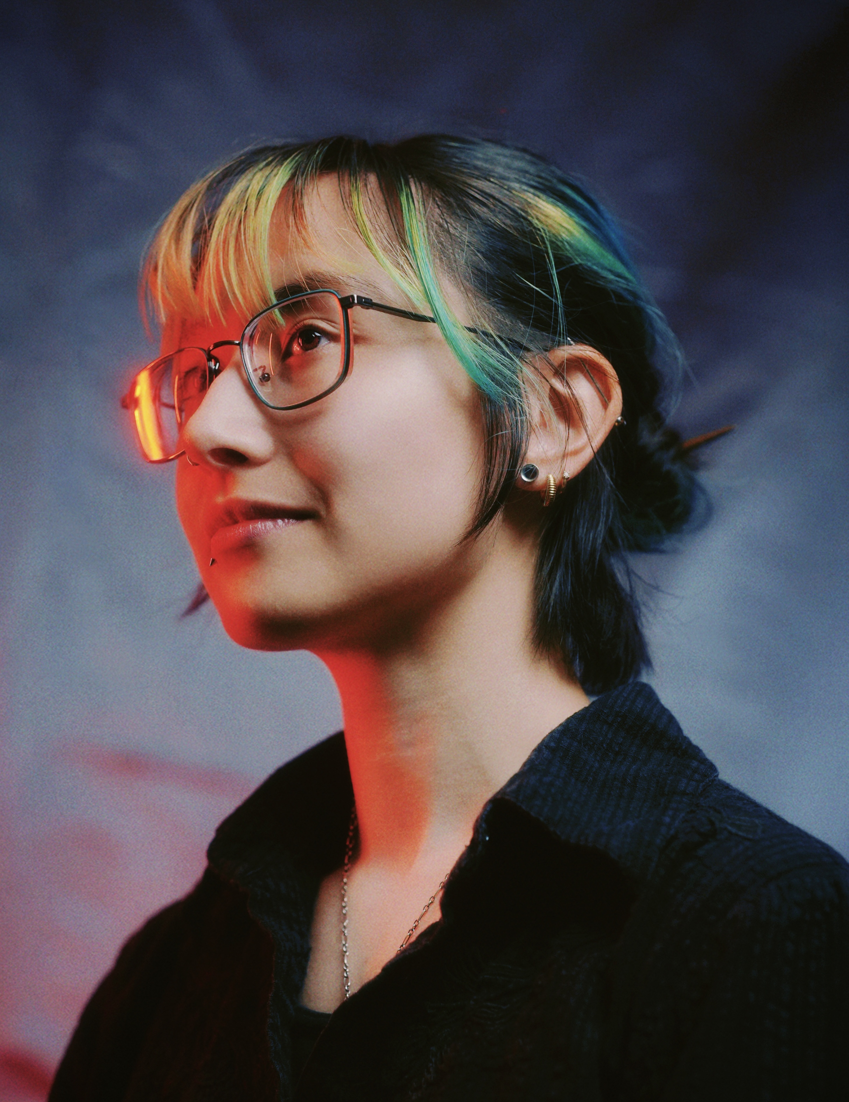 Three quarter profile of Wraven Watanabe facing left, looking up and beyond the camera. 
     Dark hair is held back in a bun, with loose green bangs. Orange light reflects off of Wraven's glasses.